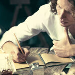 Writer at work. Handsome young writer sitting at the table and writing something in his sketchpad