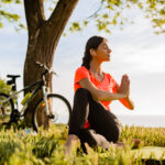 slim beautiful woman doing sports in morning in park doing yoga on mat in colorful fitness outfit in nature, smiling happy healthy lifestyle, calm meditation, bicycle on background
